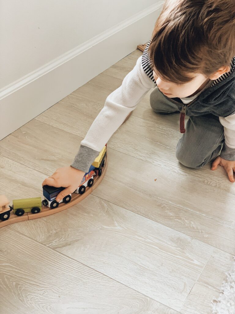 Young boy playing with a colored wooden train set on a light weathered wood floor